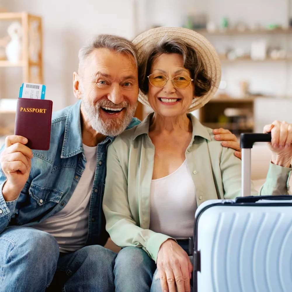 Casal mais velho sorrindo no sofá de casa, um deles segurando o passaporte com os cartões de embarque